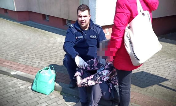 policjant kuca przy kobiecie siedzącej na chodniku, obok nich stoi druga kobieta