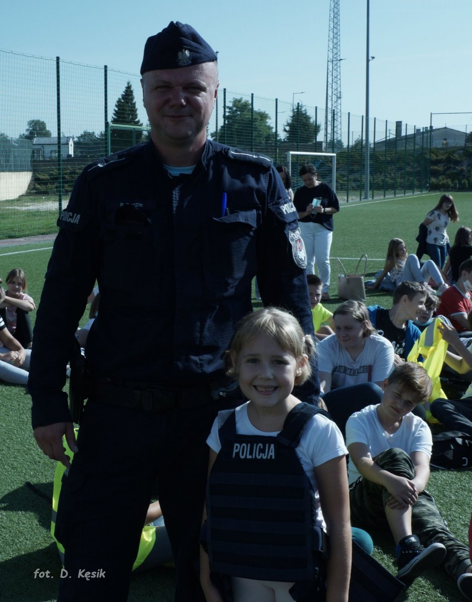 Policjant w mundurze z dziewczynką na boisku w tle dzieci siedzą na trawie